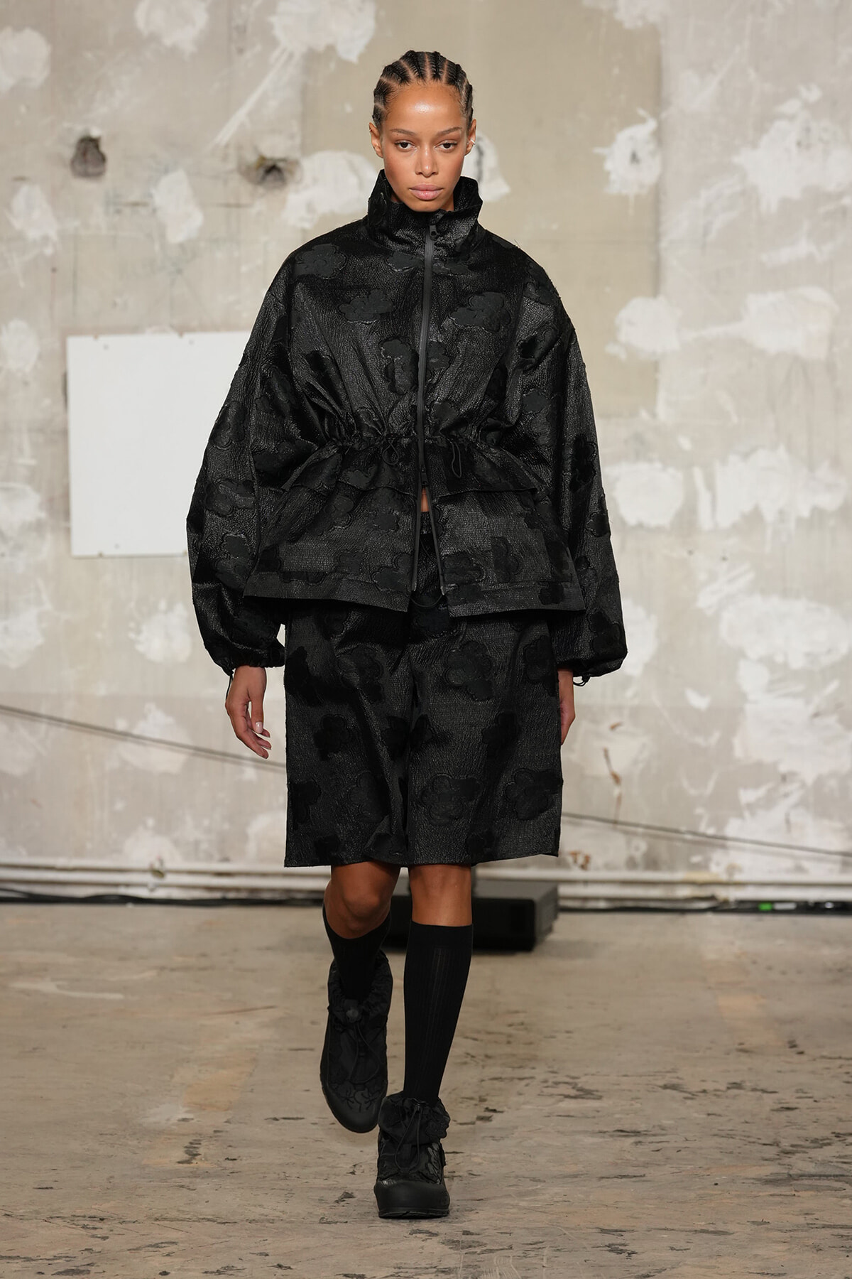 Model walking a runway in a black, textured floral set: oversized zip-front jacket cinched at the waist and matching knee-length shorts with black socks and boots.
