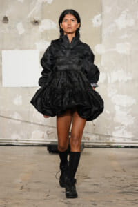 Model walking a runway in a black sculptural dress with a voluminous, tiered skirt and braided hair.