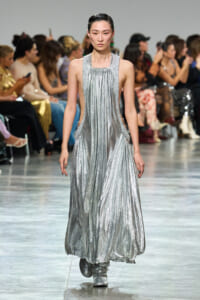 Female model walks a silver metallic gown down a runway with a seated audience on both sides of the aisle.