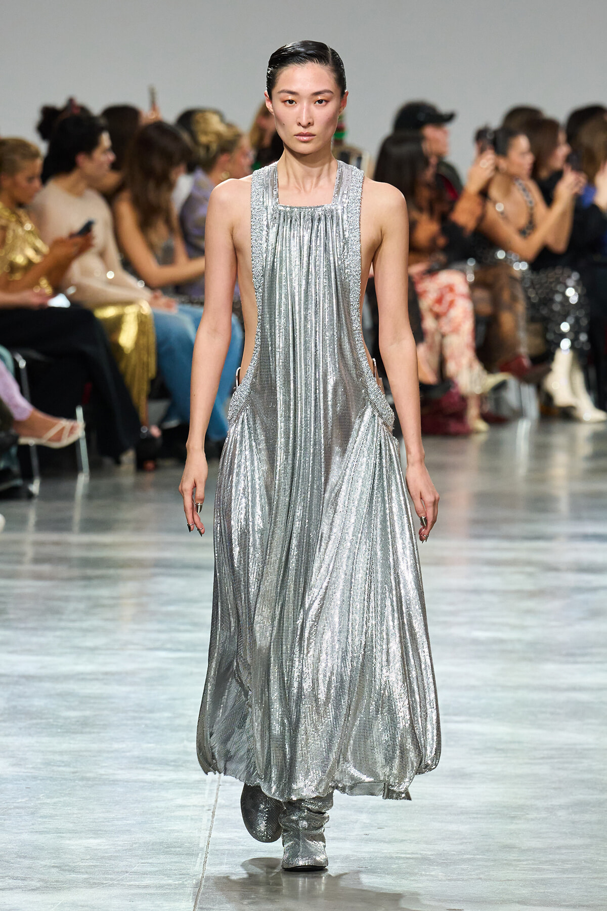 Female model walks a silver metallic gown down a runway with a seated audience on both sides of the aisle.