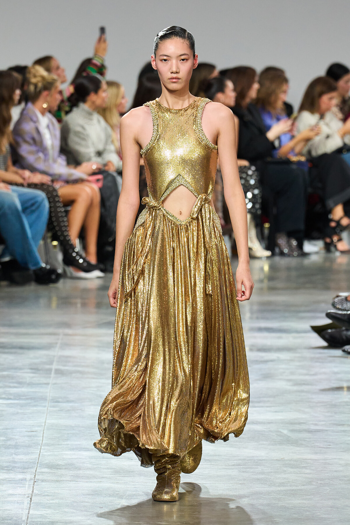 Model walks the runway in a gold metallic halter dress with a cutout at the waist and a flowing pleated skirt.