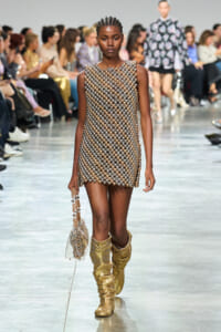 Model walking a runway in a silver-gold beaded mini dress, matching beaded handbag, and slouchy gold boots on a fashion show catwalk.