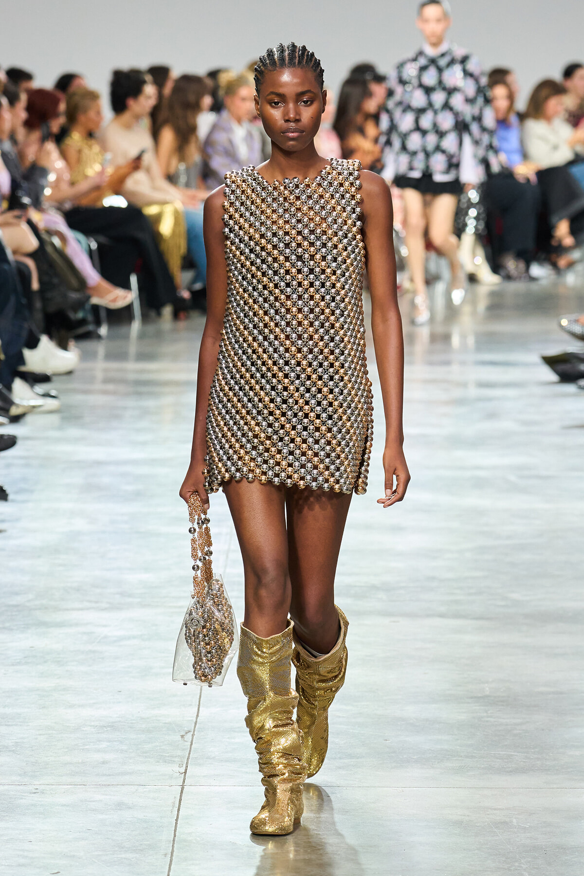 Model walking a runway in a silver-gold beaded mini dress, matching beaded handbag, and slouchy gold boots on a fashion show catwalk.