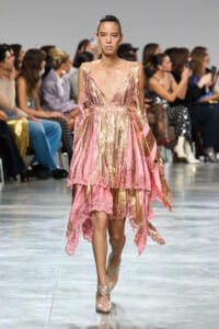 Model walking a pink and gold metallic gown with lace trim on a fashion runway