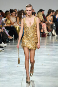 Model walks a fashion show wearing a gold textured mini dress, carrying a small matching gold bag with chain strap.