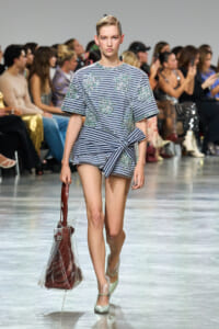 Model walks the runway in a blue striped dress with teal floral embroidery and a front knot, carrying a clear tote.