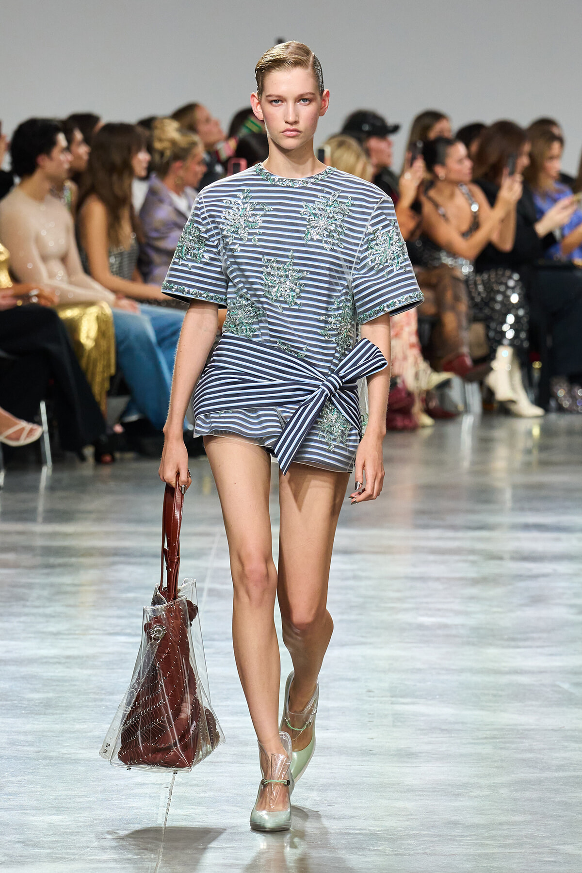 Model walks the runway in a blue striped dress with teal floral embroidery and a front knot, carrying a clear tote.