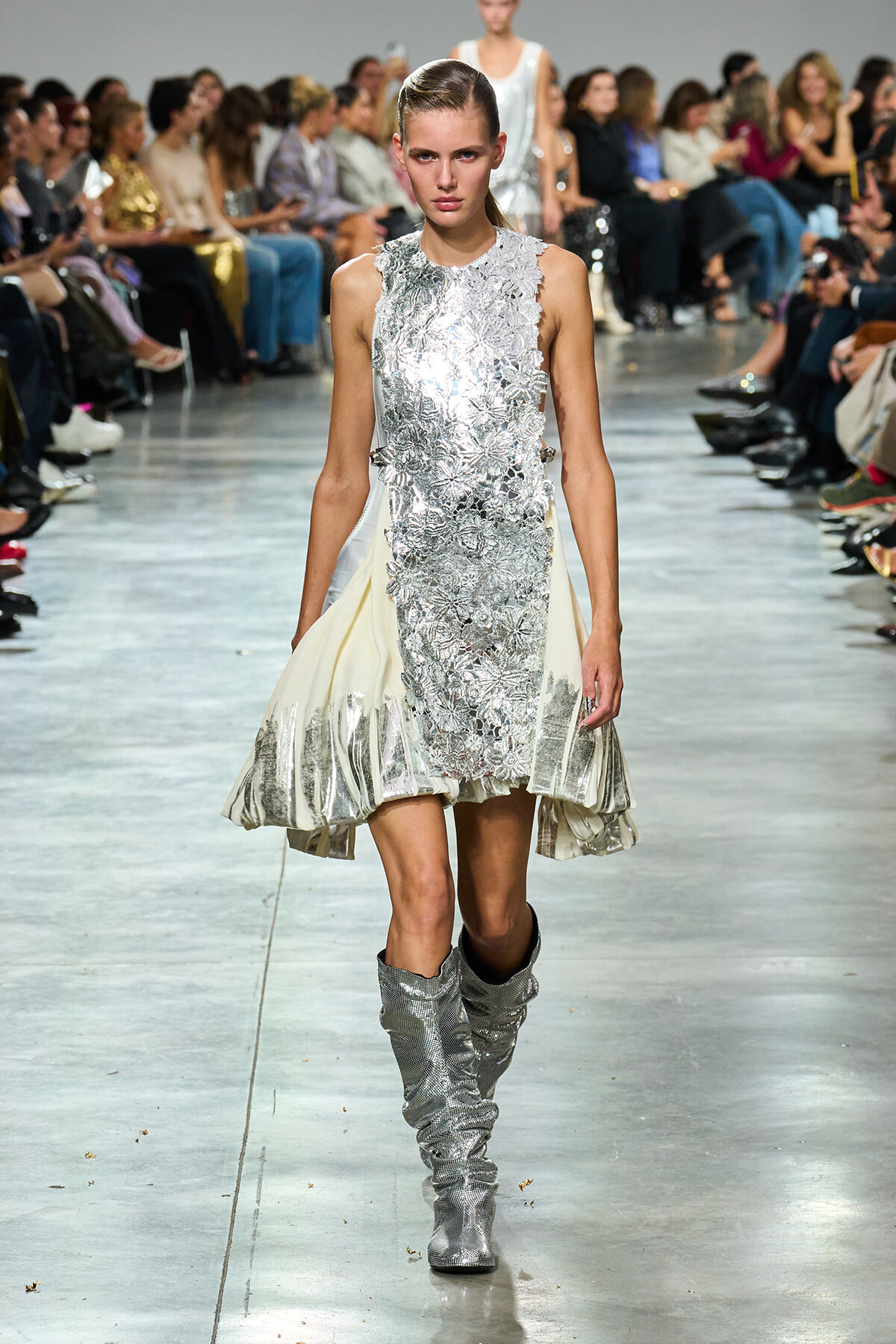 Model walking the runway in a silver lace-embellished dress with side panels and matching metallic knee-high boots.