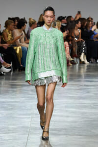 Model walks the runway in a mint green oversized knit sweater layered over a lace-trimmed dress; audience watches from the sidelines elsewhere in the venue.
