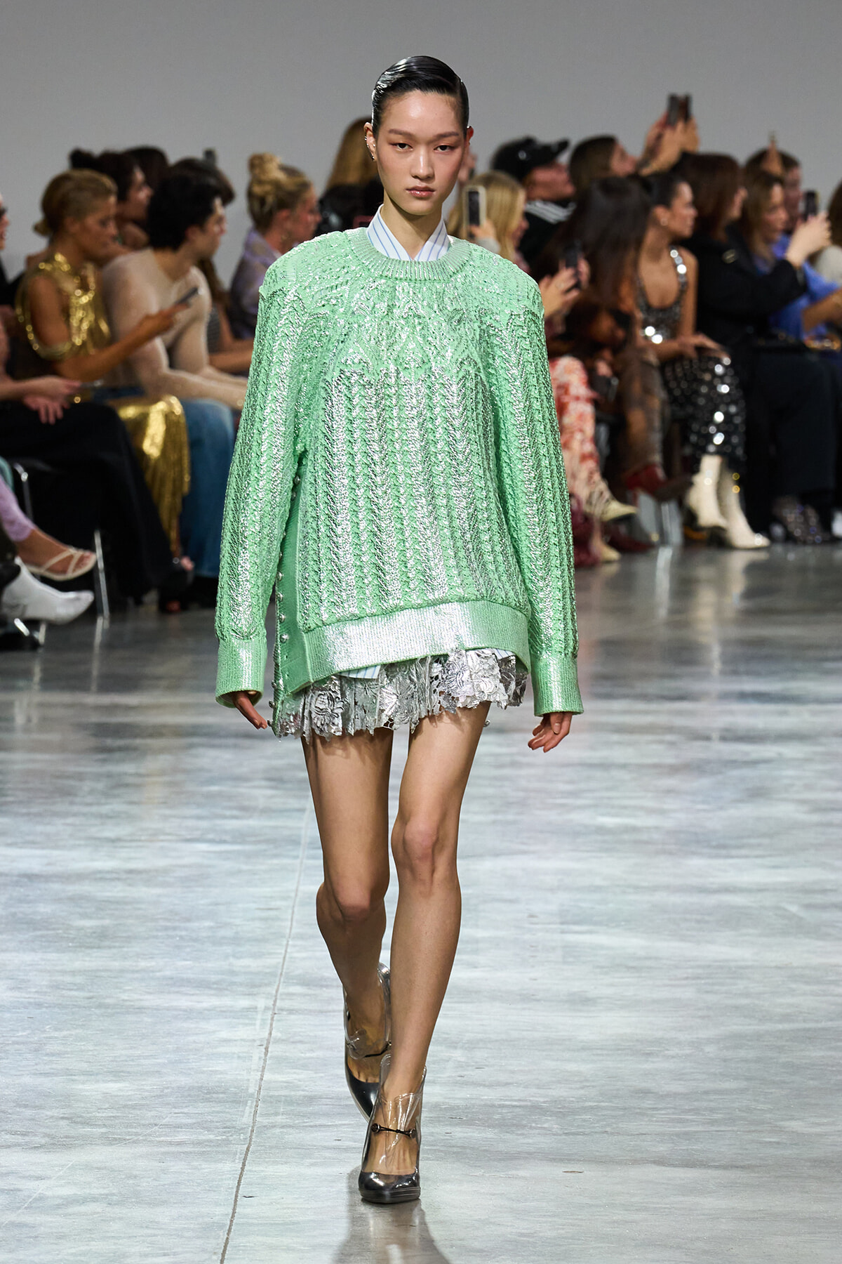 Model walks the runway in a mint green oversized knit sweater layered over a lace-trimmed dress; audience watches from the sidelines elsewhere in the venue.