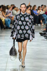 Model walking a runway in a black floral sweater over a striped shirt, black pleated skirt, silver ankle boots, and a clear bag with dark interior.