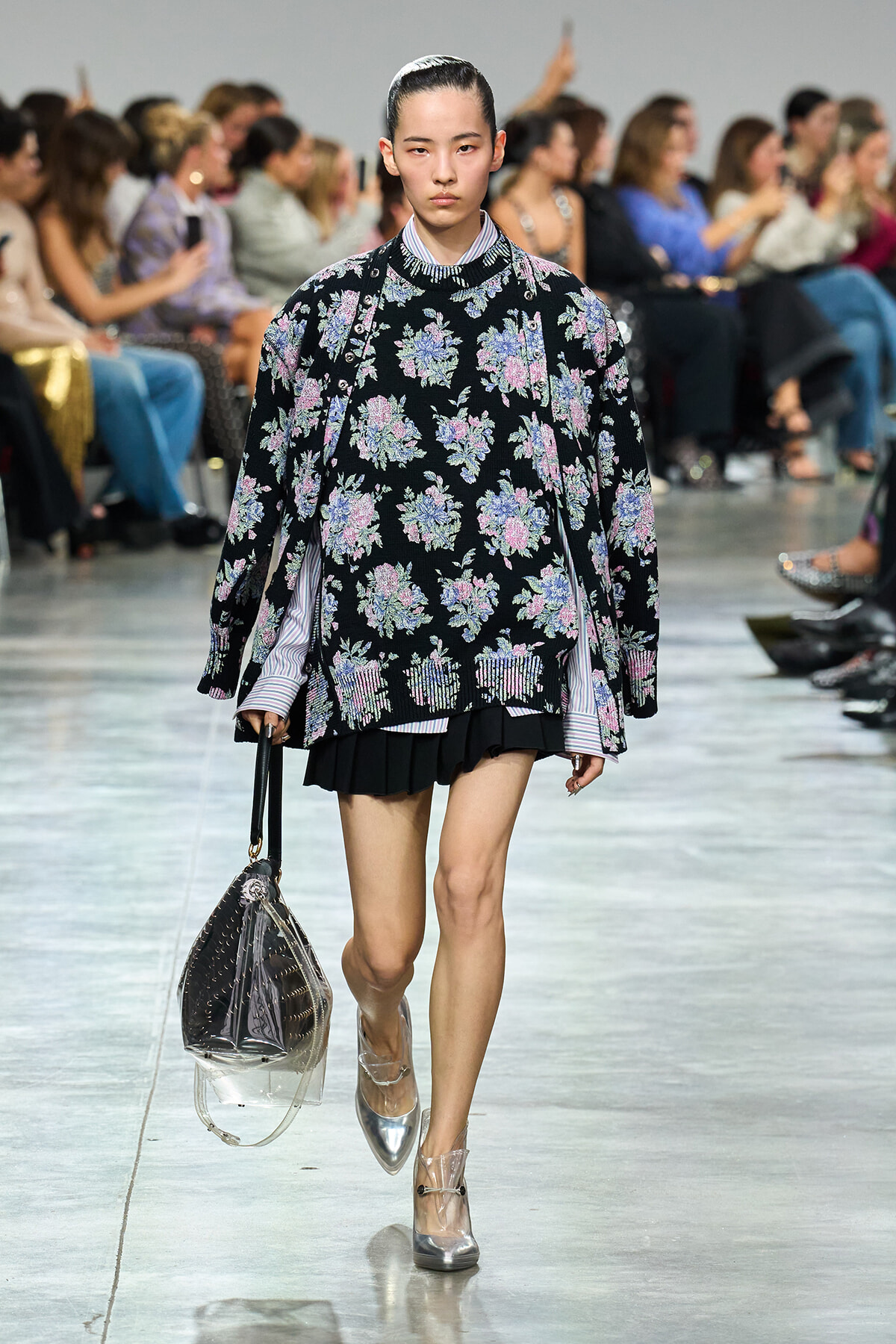 Model walking a runway in a black floral sweater over a striped shirt, black pleated skirt, silver ankle boots, and a clear bag with dark interior.