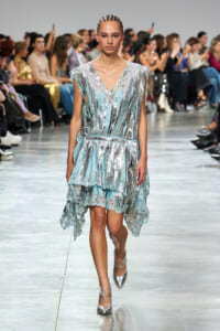Model walks a shimmering blue and silver lace dress down a runway as the audience watches the show.