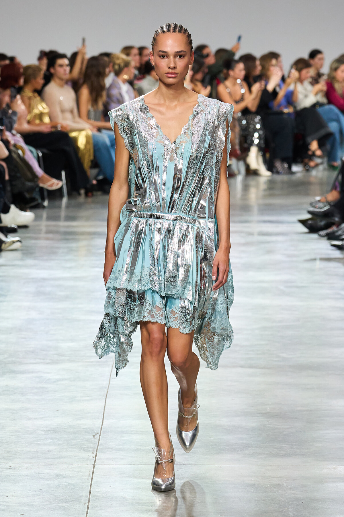 Model walks a shimmering blue and silver lace dress down a runway as the audience watches the show.