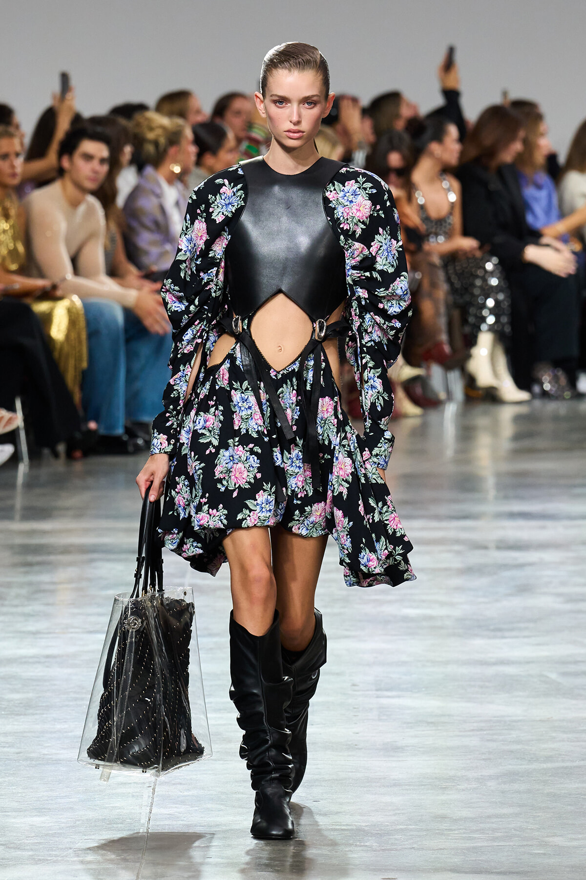 Model walks the runway in a black floral print dress with cutouts and puffed sleeves, paired with knee-high black boots, audience in background.