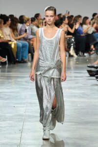 Runway model in a silver sequined sleeveless top and draped metallic skirt with a knot at the hip, wearing white boots in a fashion show.