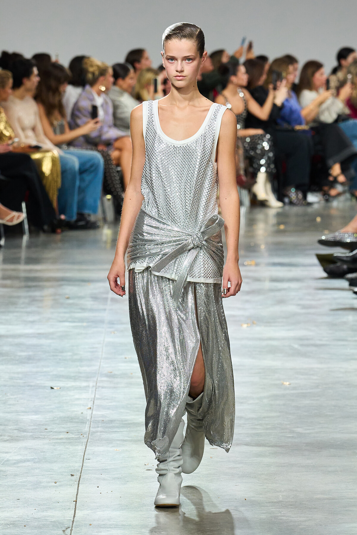 Runway model in a silver sequined sleeveless top and draped metallic skirt with a knot at the hip, wearing white boots in a fashion show.