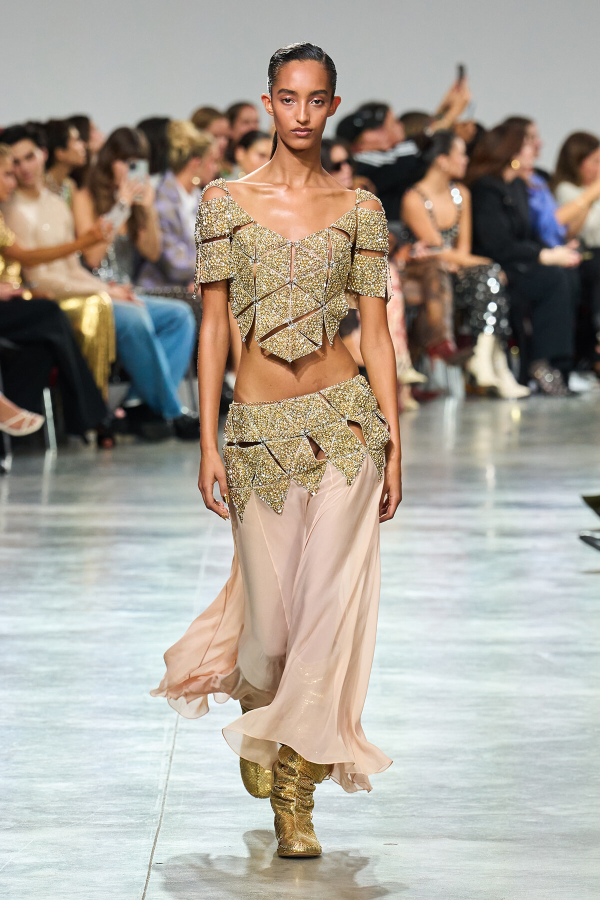 Model walks a runway in a gold geometric, beaded two-piece outfit with off-shoulder sleeves and a pale pink flowing skirt, gold boots visible.