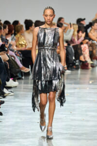 Model walks a metallic silver and black tiered dress down a runway, hands in pockets, spectators in the background.
