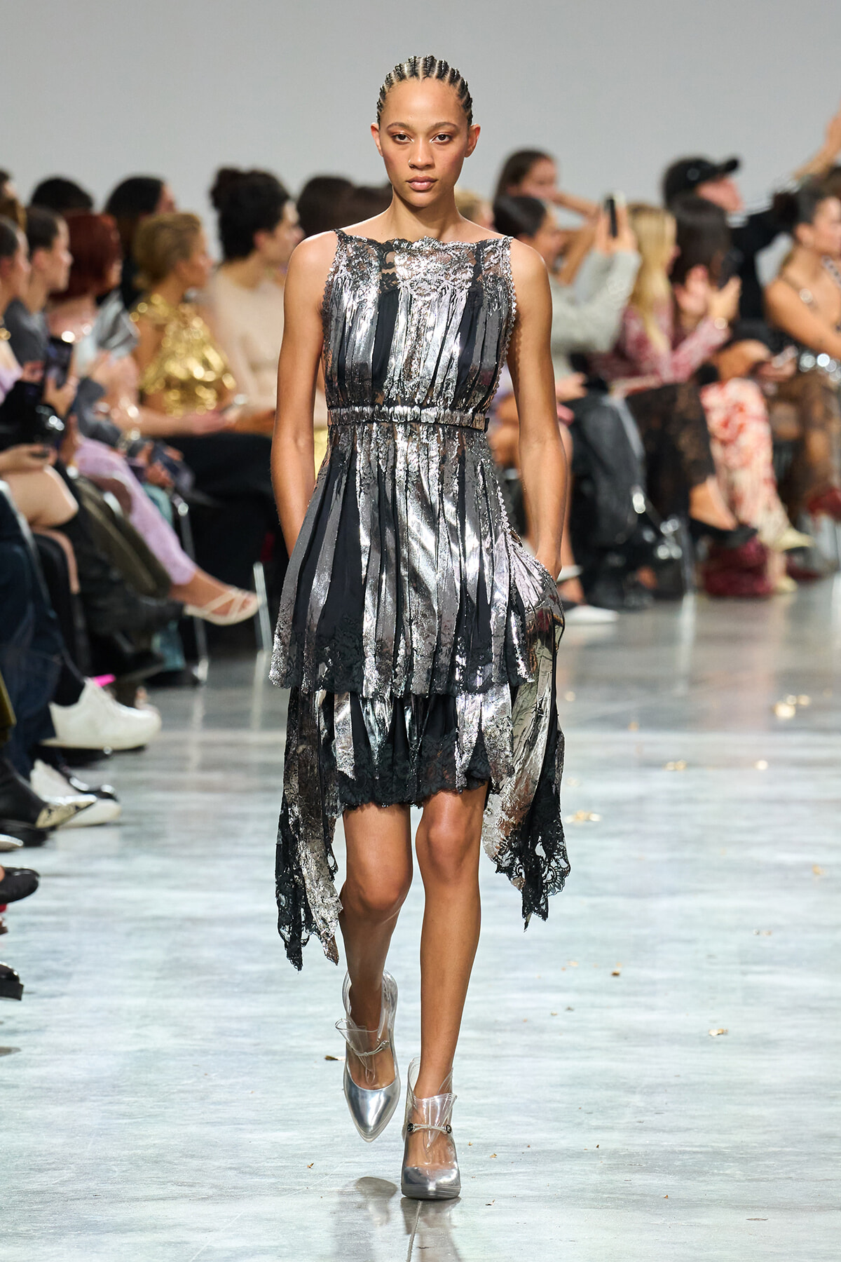 Model walks a metallic silver and black tiered dress down a runway, hands in pockets, spectators in the background.