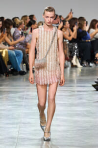 Model walks the runway in a pale pink, beaded mini dress with circular discs and a silver chain crossbody bag; metallic heels complete the look.