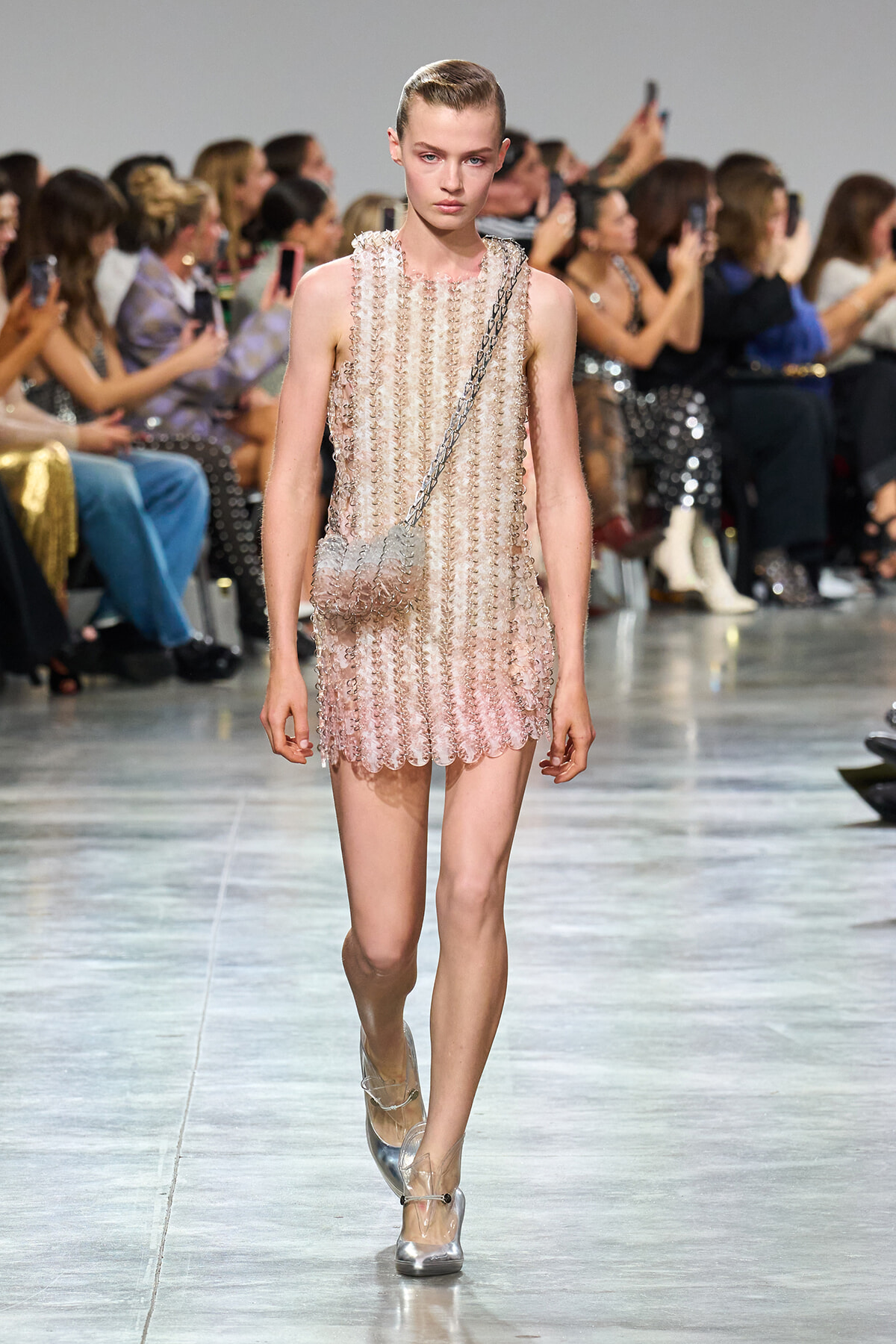 Model walks the runway in a pale pink, beaded mini dress with circular discs and a silver chain crossbody bag; metallic heels complete the look.