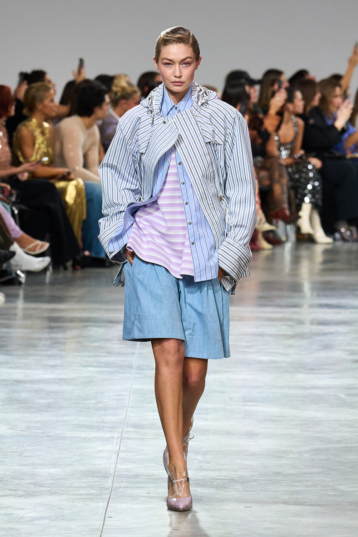Model walking a runway in oversized striped shirt over lavender top and light blue shorts; audience watches from the sides.
