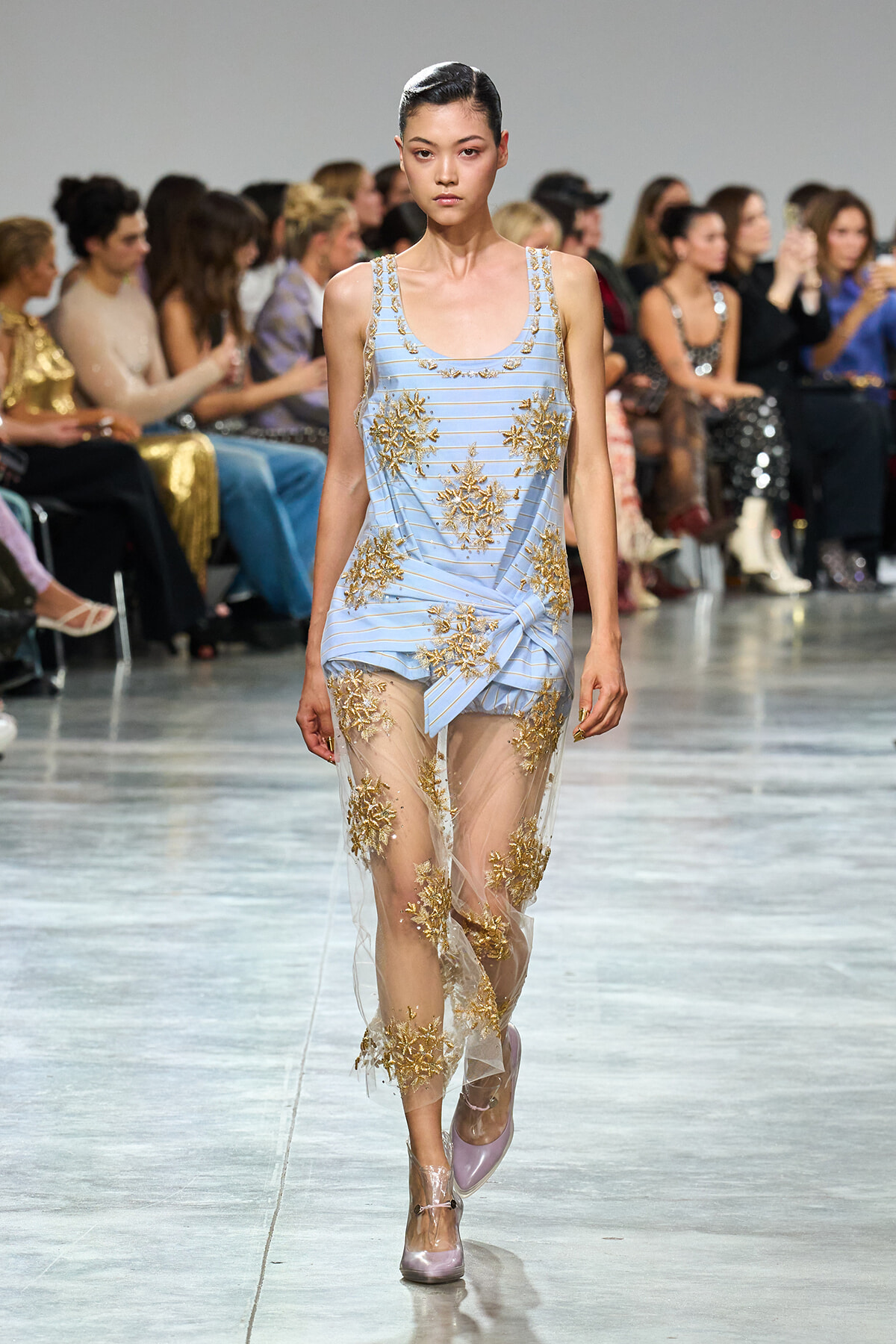 Model walks the runway in a light blue striped dress with gold floral embroidery and a sheer, embroidered overlay trailing to the floor. Pink heels complete the look.
