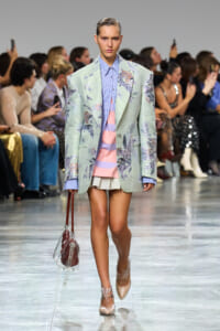 Model walking a fashion runway in a mint floral blazer over a pastel striped shirt and a beige mini skirt, carrying a clear bag.