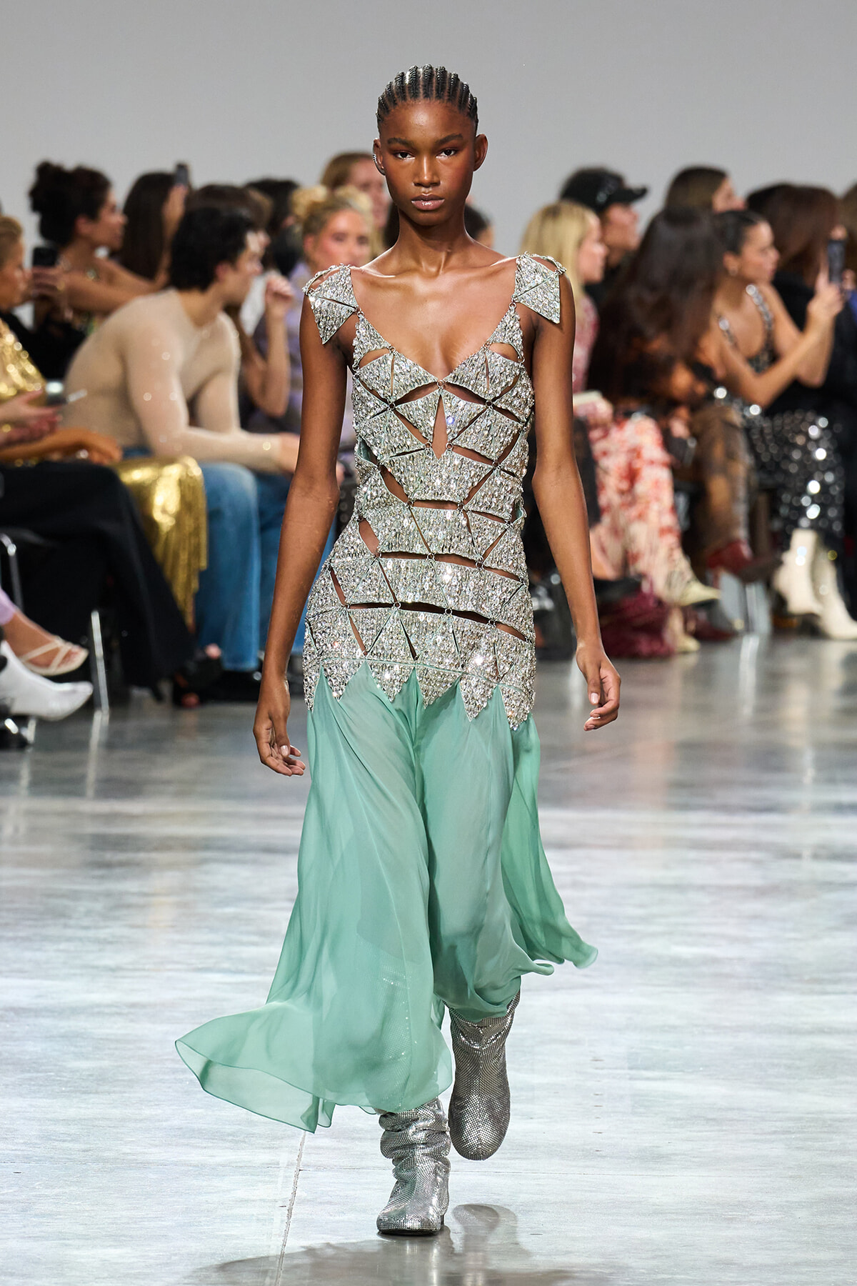 Model walks the runway in a sparkling silver lattice bodice with mint green flowing pants and silver boots; audience blurred in the background.