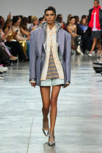 Model walking the runway in an oversized lavender coat over a peach striped shirt and mint shorts; audience in background.