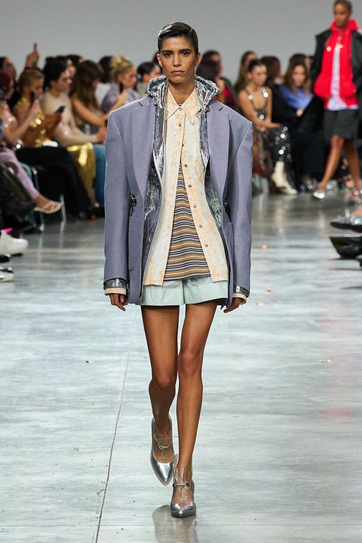 Model walking the runway in an oversized lavender coat over a peach striped shirt and mint shorts; audience in background.
