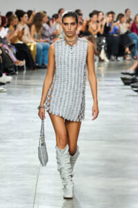 Model walks a runway in a silver chainmail sleeveless mini dress with a scalloped hem, paired with white knee-high boots and a matching chain-link handbag.