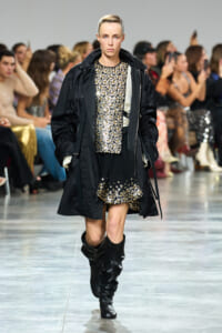 Model walks the runway in a black coat over a sequined dress with silver embellishments and black boots as the audience watches behind him/her.