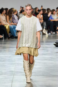 Model walking a fashion runway in a cream knit sleeveless sweater over a white tee, with a beige bubble skirt and tall beige boots. Audience in background