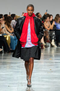 Runway model in a bright red hoodie under a black leather coat, wearing striped shorts and clear heels as spectators watch the show.