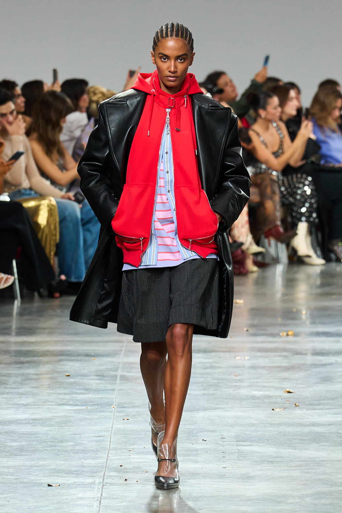 Runway model in a bright red hoodie under a black leather coat, wearing striped shorts and clear heels as spectators watch the show.