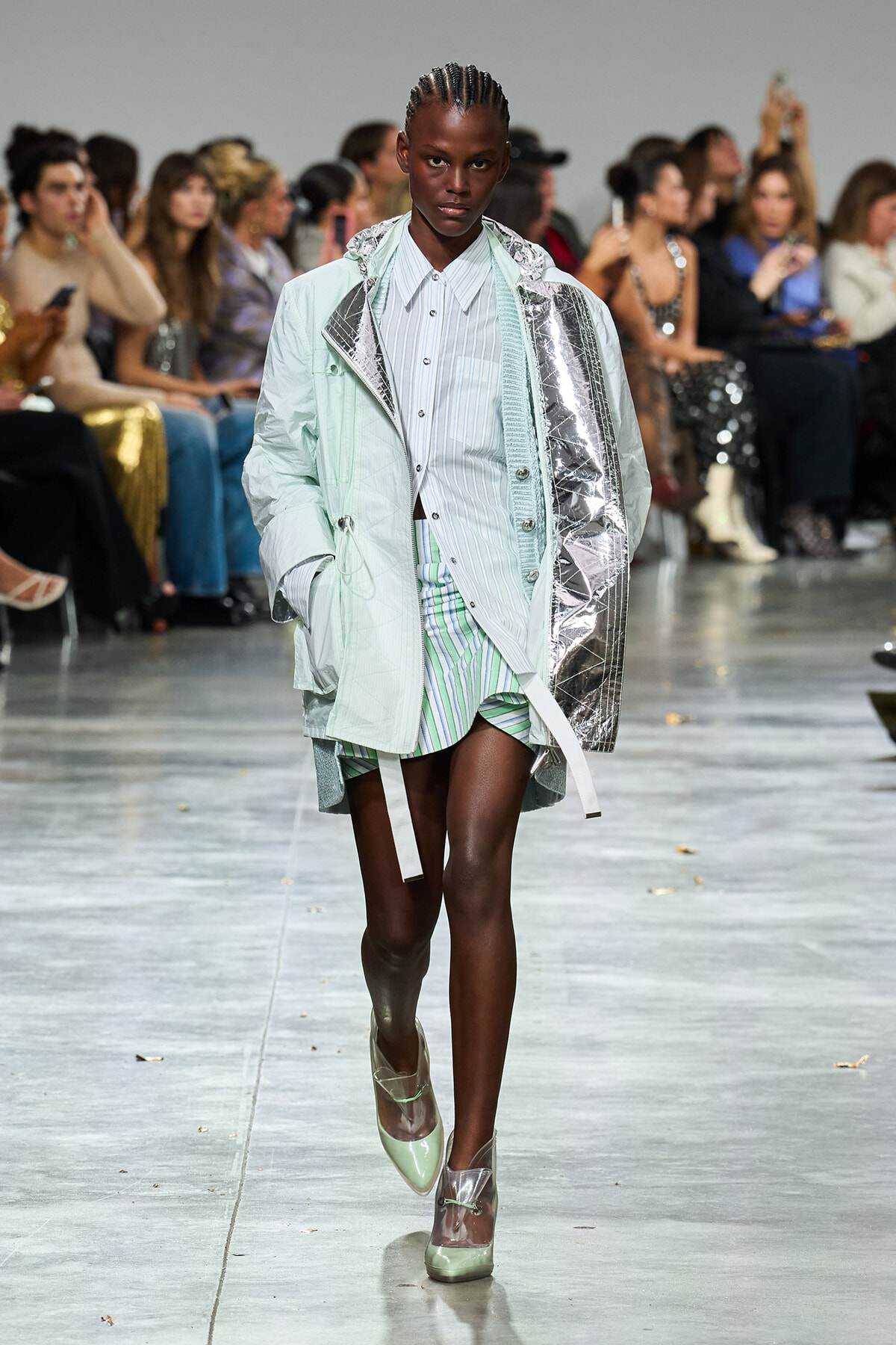 Runway model in a pastel green oversized coat with metallic inner panel, striped shirt, and striped shorts; audience seated along the runway.