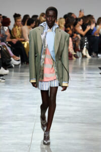 Runway model wearing an oversized sage blazer over pastel color blocks (light blue shirt, pink stripes, blue striped shorts) with silver ankle boots.