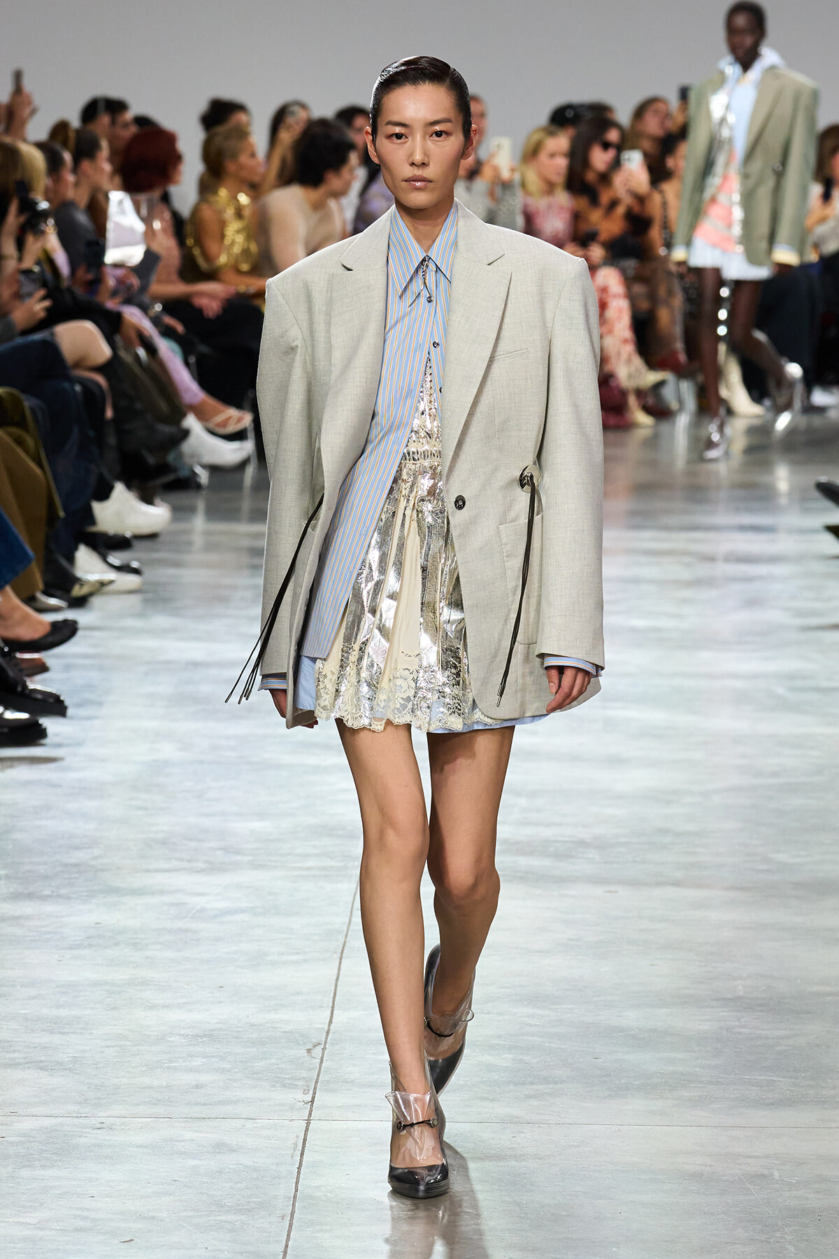 Model walks the runway in an oversized light-gray blazer over a blue striped shirt and metallic silver lace mini skirt; audience watches from the sides.