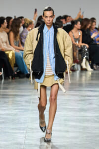 Model walks down a runway wearing a tan oversized jacket over a blue striped shirt and beige mini skirt, with audience in the background.
