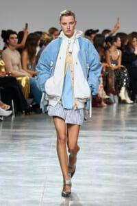 Model walking a fashion runway in an oversized light-blue jacket layered over a white vest and pastel-striped shorts, with an seated audience in the background.