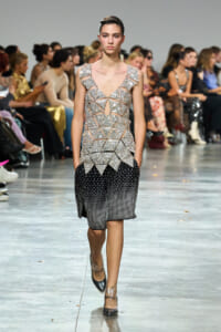 Model walks the runway in a silver geometric sequined top and black dotted shorts, with spectators in the background on a fashion show runway.