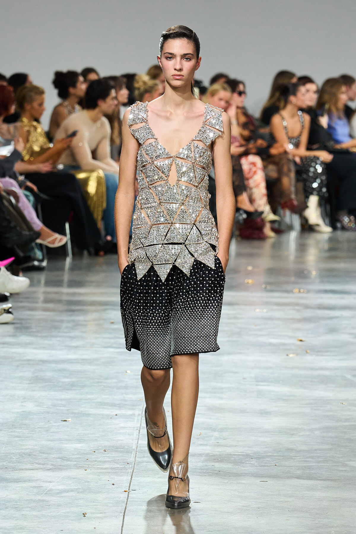 Model walks the runway in a silver geometric sequined top and black dotted shorts, with spectators in the background on a fashion show runway.