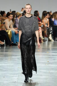 Model walks the runway in a shimmering black wrap skirt and a dotted short-sleeve top, audience in the background.