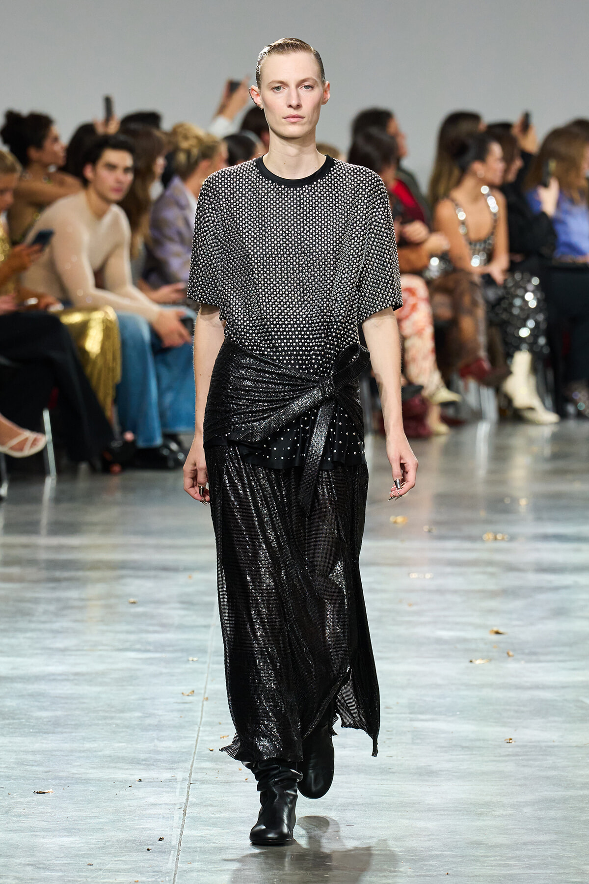Model walks the runway in a shimmering black wrap skirt and a dotted short-sleeve top, audience in the background.