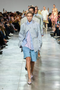 Model walks the runway in an oversized blue striped jacket over a lavender striped shirt and light-blue shorts; audience and photographers line the sides.
