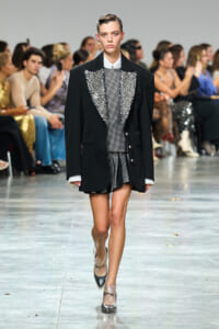 Model walks the runway in a black blazer over a shimmering jeweled outfit with silver studs; audience in background watches.