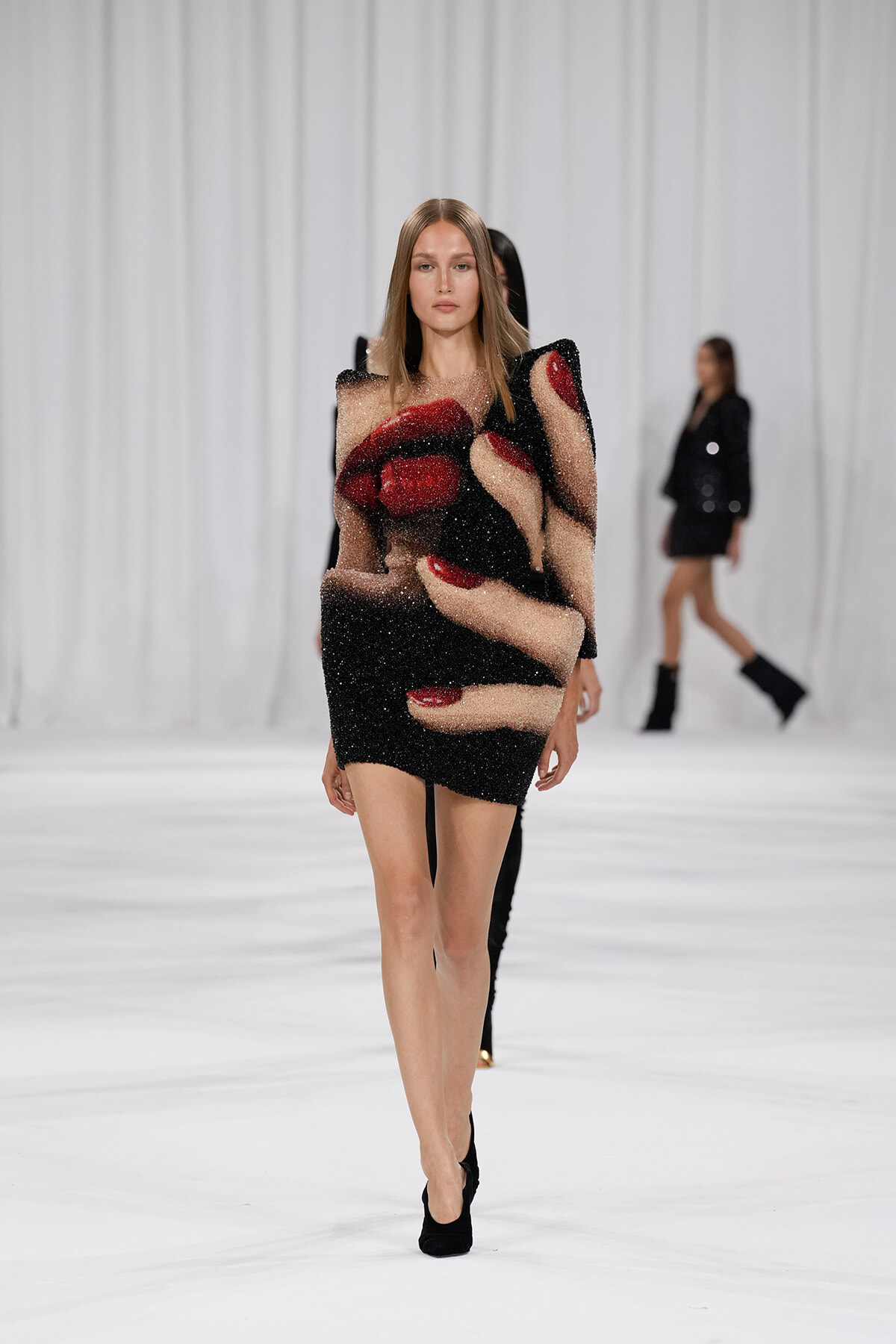 Model walking the runway in a sparkly black mini dress featuring oversized nude-colored hands and red nails against a white backdrop.