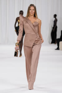 Model walks the runway in a blush-pink, one-shoulder suit with pleated detailing and matching wide-leg trousers, carrying a small rectangular handbag.